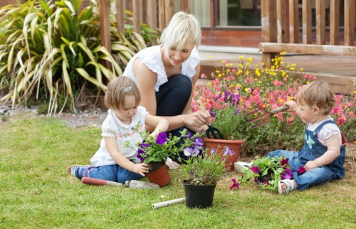 In a well-maintained garden, a smiling woman with light blonde hair sits on a lush green lawn alongside two young children. The woman is engaged in gardening activities, holding a small trowel and tending to flowering plants in various pots, with one pot containing vibrant purple flowers and another with pink blooms. The children, both dressed in casual summer clothing, are sitting on the grass, each with a small plant pot filled with colorful flowers, actively participating in planting or watering. The garden features a flower bed with bright pink and yellow blossoms adjacent to a wooden deck area, which is partially visible in the background. Surrounding the scene are tall, leafy plants with variegated foliage, providing a natural border to the outdoor space. The environment appears to be outdoors on a sunny day with clear weather, showcasing a peaceful, family-friendly gardening setting that demonstrates garden care, planting activities, and outdoor leisure, reflecting the services offered by Landscapers Islington for garden maintenance and improvement in the local area.