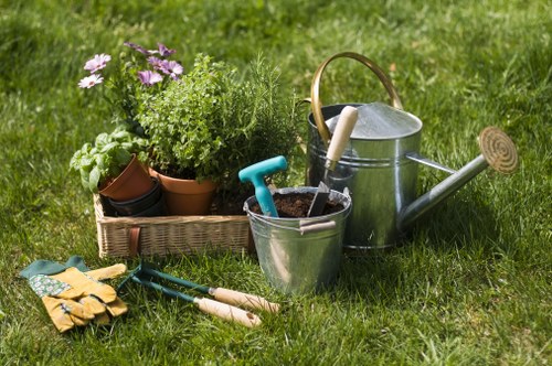 A neatly maintained backyard garden with a well-kept grass lawn featuring lush, green grass with slight variations in tone, bordered by a flower bed or shrub border not visible in the image. In the foreground, there are gardening tools including a pair of yellow gardening gloves, small hand trowels with wooden handles, and a pair of pruning shears. To the side, a wooden basket contains potted plants with green foliage and purple flowering plants, alongside a small terracotta pot. Behind the tools, there is a metal watering can with a wooden handle and a garden rake. The outdoor space is well-lit under natural daylight, suggesting clear weather, with the scene highlighting typical gardening activities and outdoor maintenance elements that align with services offered by Landscapers Islington in the area around N1 postcode. The overall setting depicts an organized, cared-for garden area suitable for lawn care and planting, reflecting professional gardening practices and outdoor landscaping efforts.