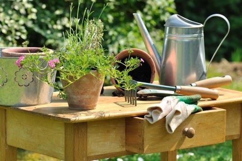 A wooden outdoor work table in a garden, displaying various gardening tools and supplies, including a galvanized watering can, terracotta pots with green plants and pink flowers, a pair of gardening gloves, a rake, and a trowel. The background features lush green foliage, suggesting a well-maintained backyard or garden space. The scene captures a moment of garden maintenance or planting, highlighting the tools commonly used in lawn care and planting activities. The natural daylight and outdoor setting imply a bright, clear day, with the scene emphasizing organized gardening preparation and care typical of professional landscaping services in Islington or surrounding areas. The overall arrangement and elements reflect an active gardening environment, suitable for garden renovation projects or outdoor space improvements by Landscapers Islington.