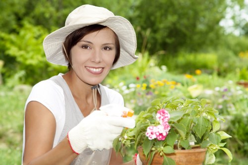 A woman wearing a white sun hat, white gardening gloves, and a light grey t-shirt is standing outdoors in a landscaped garden in Islington, tending to a potted flowering plant with pink and white blossoms. The garden features a lush green lawn, with dense foliage and various flowering plants in the background, including shrubs and small trees. The setting appears to be a well-maintained front or back garden with a mix of natural grass, soil beds, and decorative plantings. The weather is bright and sunny, casting natural light across the scene, highlighting the vibrant colours of the flowers and greenery. This image reflects professional gardening and landscaping activities typical of local garden maintenance services, demonstrating detailed plant care and outdoor environment management in an urban setting like Islington, supporting the services offered by Landscapers Islington.