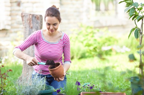 A woman with dark hair tied back, wearing a pink and white striped long-sleeve top, is tending to potted plants in a garden. She is holding a terracotta pot filled with dark soil in one hand and using a small gardening trowel in the other to transfer soil or plant material into the pot. The garden features a lush green lawn, various flowering plants, and a partially visible wooden bench in the background. Surrounding the woman are additional potted plants and garden tools, with a blurred backdrop of trees and a brick wall, indicating a well-maintained outdoor space suitable for gardening services in Islington. The scene appears to be during daylight hours on a mild, overcast day, emphasizing a natural outdoor environment with a focus on gardening activities and plant care.