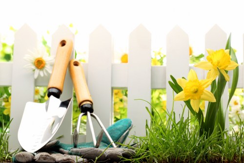 The image depicts a garden scene with a white picket fence as a backdrop, illuminated by natural daylight, suggesting a bright and sunny weather condition. In the foreground, there's a patch of well-maintained, lush green grass with a dense and even coverage, typical of lawn areas often included in residential garden designs in Islington. To the right, a small bed of vibrant yellow daffodils with tall, green leaves adds a splash of colour, contrasting with the greenery and contributing to the garden's natural aesthetic. On the left, gardening tools—two trowels with wooden handles and a hand fork, positioned upright and leaning against each other—are placed on a dark cloth or mat, indicating active gardening or recent planting activities. The background features the white wooden fence with slightly pointed tops, beyond which blurred hints of additional garden plants and foliage are faintly visible, suggesting a well-tended outdoor space. The scene captures a peaceful, manageable garden environment suitable for outdoor maintenance services like lawn care, planting, and general garden landscaping, typical of residential properties in the London Borough of Islington, aligning with gardening service offerings from Landscapers Islington aimed at enhancing outdoor curb appeal.