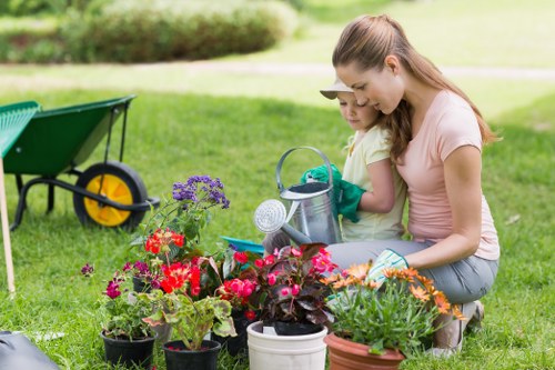 In a well-maintained garden, a woman and a young girl are engaged in planting and caring for a variety of flowering plants and foliage in pots arranged on the lush green lawn. The garden features a neatly trimmed hedge and a backdrop of a grassy area, with a wheelbarrow positioned nearby for gardening tasks. The soil appears rich and well-prepared, and the flowering plants display vibrant reds, purples, and oranges, creating a colourful scene suitable for outdoor gardening activities. Both are dressed casually for gardening, with the woman wearing short sleeves and the girl in a light T-shirt, indicating mild weather conditions. The scene captures an outdoor space designed for leisure and horticultural enjoyment, with natural light enhancing the different textures of the garden beds, containers, and grass, reflecting the services of a professional gardening company such as Landscapers Islington, who may maintain such landscaped outdoor areas in the local area of Islington, London, within postcode N1, supporting accessibility and visual harmony in the garden environment.