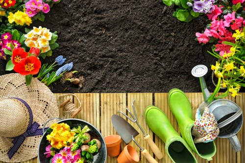 The image shows a neatly organized garden scene with a wooden decking area bordered by colourful flower beds containing pink, purple, and yellow blooms. The foreground features gardening tools including a small hand trowel, a cultivator, and a pair of bright green rubber boots placed on the decking. A woven straw garden hat with a dark ribbon rests on the edge of the decking, alongside a black plastic pot filled with freshly planted flowers, and several small terracotta pots. In the background, a patch of dark, rich soil is prepared for planting, with a few partially visible gardening gloves and watering cans nearby. The overall setting suggests an outdoor space in a residential garden, possibly in Islington or nearby, where local gardening services like Landers Islington could assist with lawn care, planting, and landscape maintenance. The scene is illuminated by natural daylight, indicating favourable weather conditions suitable for outdoor gardening activities.