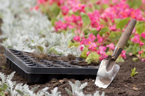 A close-up of a gardening scene in a landscaped outdoor space, featuring a black planting tray on freshly turned soil, with a small hand trowel embedded in the earth. In the background, vibrant pink and white flowering plants add colour to the garden, which also includes neatly maintained flower beds and lush green foliage. The garden has a well-defined layout with flower bed borders, and the soil appears rich and well-prepared for planting. The weather appears mild and bright, with natural light illuminating the garden environment. This setting reflects the gardening and outdoor maintenance services offered by Landscapers Islington, situated near the postcode area of N1 in London, helping keep residential outdoor spaces attractive and healthy.