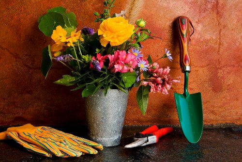 A close-up view of a garden workspace featuring a metallic flowerpot filled with a vibrant assortment of fresh garden flowers, including yellow, pink, purple, and white blooms, placed on a dark stone surface against a warm, terracotta-colored wall. Nearby, there is a pair of red-handled pruning shears, a small green gardening trowel, and a pair of yellow and green striped gardening gloves, suggesting active gardening or landscaping work. The scene highlights tools and floral arrangements typical of garden maintenance in an outdoor or greenhouse setting, with soft natural lighting emphasizing the varied textures of the flowers, foliage, and tools. This setup is representative of a professional gardening environment, suitable for services provided by companies like Landscapers Islington, promoting gardening, lawn care, and outdoor landscaping within the local area, possibly in London or nearby boroughs such as Islington or N19 postcode area.