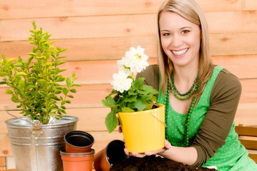 A woman with blonde hair and a cheerful expression is sitting outdoors in a garden, holding a bright yellow flower pot with a healthy, flowering plant featuring white blossoms and green foliage. Behind her, there is a background of wooden fencing or decking, suggesting a landscaped backyard area in Islington. On the table in front of her, there are various gardening items including a metal watering can, empty plant pots, and soil or compost, indicating ongoing or recent planting activity. The garden space appears well-maintained with a lush, green grassy area visible in the foreground, and the overall scene is lit by natural daylight, suggesting clear weather. This setting reflects typical gardening and landscaping activities provided by local companies like Landscapers Islington, focused on garden design, plant care, and outdoor maintenance services.
