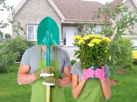 In a front garden with a well-maintained lawn and lush green shrubs, two individuals are engaged in gardening activities. One person, wearing a green apron, gloves, and a gardening helmet with a face shield, is holding a garden shovel upright. The other, dressed in a grey t-shirt, pink gloves, and a pink apron, is holding a bright yellow flowering plant together with green foliage in front of their face. Behind them, there is a neatly trimmed hedge, a flower bed with small plants, and a background of a residential house featuring beige brick walls, white-framed windows, and a grey tiled roof. The garden environment appears clean, orderly, and suitable for outdoor maintenance or planting projects. The weather looks mild with natural daylight illuminating the scene, consistent with typical gardening conditions in Islington, London. This image reflects professional gardening and landscaping services offered by Landscapers Islington, emphasizing garden landscaping, planting, and outdoor yard enhancements commonly provided in urban gardens within the postcode area.