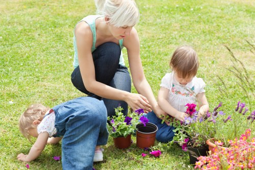 A gardener, likely a woman with short blonde hair wearing a light blue sleeveless top and dark jeans, is kneeling on a well-maintained grassy lawn within an outdoor garden space in Islington. She is actively tending to a flower bed, where two young children are also engaged; one child, dressed in light clothing, is kneeling and reaching into the soil, while the other, wearing a white top with pink flowers, is sitting nearby holding a small pink flower. The flower bed contains various colourful flowers with purple, pink, and orange blooms, bordered by dark soil. Nearby, there are flower pots and gardening tools, indicating ongoing planting activity. The background features a neatly trimmed hedge or shrubbery, with a clean, lush lawn area extending behind the scene. The weather appears bright and clear, providing natural lighting that highlights the vibrant colours of the flowers and plants, illustrating a typical daytime gardening or outdoor family activity in an urban garden setting, in line with landscaping services offered by Landscapers Islington.