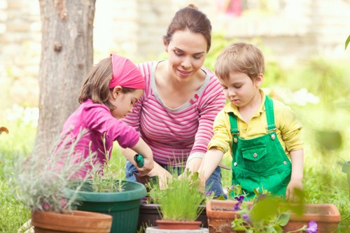 A woman and two children, a girl and a boy, are engaged in gardening activities in an outdoor garden space. The woman, dressed in a pink and white striped top, is leaning over and smiling as she assists the girl, who is wearing a pink headband and a purple long-sleeved shirt, with planting in a large green pot. The boy, wearing a yellow shirt with green overalls, stands nearby observing the activity. The garden features a variety of potted plants on a wooden surface, with some plants showing fresh green foliage and purple flowers. In the background, there is a mature tree with textured bark, a wooden fence, and lush green grass and foliage, indicating a well-maintained outdoor space typical for residential gardening in the Islington area. The scene is illuminated by natural daylight, suggesting a clear weather day suitable for outdoor gardening activities, reflecting the careful attention to garden maintenance and plant care supported by local gardening services such as Landscapers Islington.