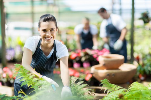 A young woman with dark hair tied back, smiling, and wearing a striped apron and gardening gloves, is tending to a lush green garden bed with vibrant ferns and other foliage. She is positioned in the foreground, leaning slightly forward with both hands on the plants, appearing engaged in gardening work. In the background, slightly out of focus, there are two other individuals wearing aprons and gloves, actively working among potted flowering plants and garden containers, on a bright and sunny day inside a greenhouse environment with glass panels allowing natural light. The scene features a well-maintained outdoor garden area with healthy plants, textured soil beds, and decorative water features, illustrating a professional gardening or landscaping service setup relevant to local outdoor maintenance and gardening care in Islington.