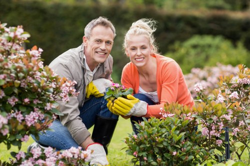A man and a woman are kneeling together in a well-maintained garden with lush green grass and flowering shrubs, including pink and white blossoms. The man, dressed in a light grey shirt, and the woman, wearing an orange sweater, are smiling at the camera while holding a flowering plant between them. They are wearing gardening gloves, indicating they have been working on maintaining or planting the garden. In the background, there are additional green bushy plants and trees, with sunlight illuminating the outdoor space, suggesting a bright, clear day. This scene depicts a typical landscaped garden area suitable for gardening and outdoor maintenance services offered by companies like Landscapers Islington, located in London. The overall environment shows a tidy garden with a healthy lawn, flowering plants in bloom, and a natural outdoor setting conducive to gardening activity and enhancement.